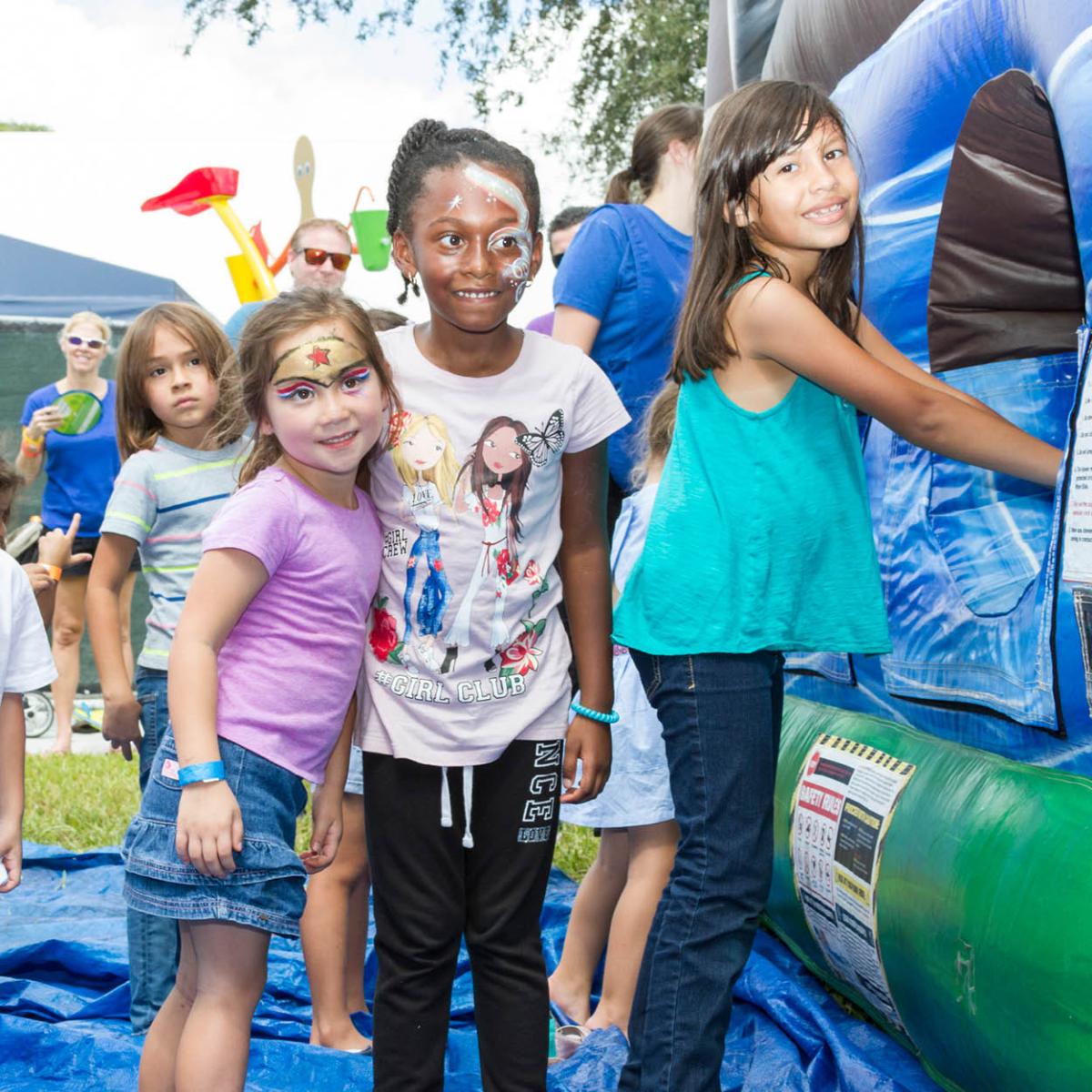 A group of children enjoying a fun outdoor event, with face painting and a bounce house in the background.
